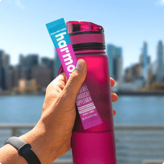 A hand holding a pink water bottle and an Advocare Harmony stick pack with a city skyline blurred in the background.