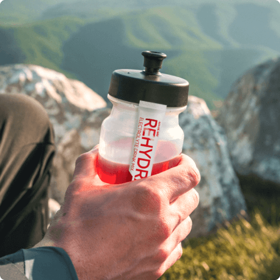 A hand holding a clear water bottle and an AdvoCare Rehydrate stick pack with hills blurred in the background.