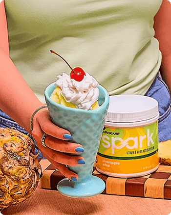 Person holding a Pineapple Whip Frosted Soft Serve in a ceramic cone next to a pineapple and canister of Spark Pineapple Whip on a table.