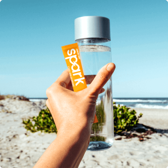 A hand holding a glass water bottle and an AdvoCare Spark stick pack with a beach blurred in the background.