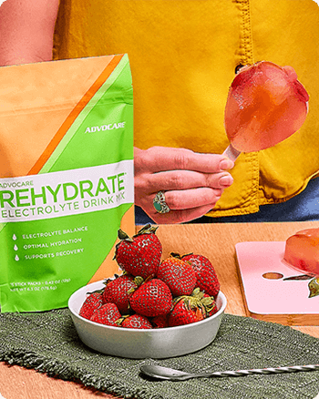 Person holding a Melonberry Popsicle with a Rehydrate Melon Stick Pack bag and bowl of strawberries on the table next to them.