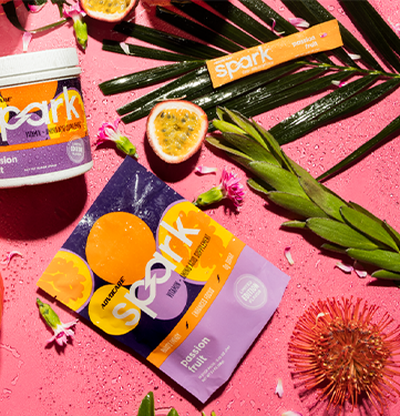 Spark Passion Fruit canister and stick pack sitting on a pink background with sliced passion fruit and floral arrangements.